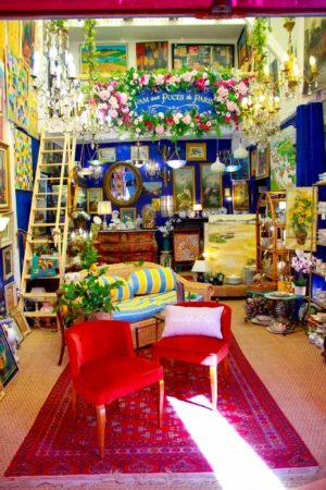 Cédric · 25/04/2026 · 1365 x 2048 Cette photographie montre l'intérieur d'une boutique ou d'un salon décoratif. Le sujet principal est un espace aménagé avec des éléments variés : meubles anciens, plantes, et objets décoratifs.La composition est symétrique, avec un tapis rouge central et des chaises disposées autour. Le cadrage inclut des étagères remplies d'articles et une décoration florale au fond. La lumière est douce et naturelle, éclairant les couleurs vives.Les couleurs dominantes sont les rouges, verts, bleus et dorés, créant une ambiance chaleureuse et festive. Le style est éclectique et chaleureux, évoquant un mélange de vintage et de convivialité.