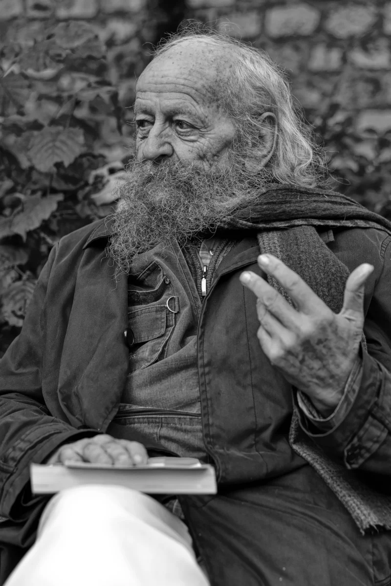 Mickaël · 11/11/2024 · 3771 x 5649 Cette photographie en noir et blanc met en scène un homme âgé au regard intense, sujet principal. Il porte une veste épaisse et une barbe blanche, gesticulant comme s'il s'exprimait avec passion.Le cadrage est serré, centré sur son visage et ses mains, avec un arrière-plan flou de pierres et de végétation. La lumière, douce et naturelle, éclaire son visage par la gauche, créant des contrastes marquants.Le style est documentaire et intime, évoquant une ambiance authentique et émouvante, presque théâtrale. Les tons gris et noirs renforcent la profondeur et la gravité de la scène.