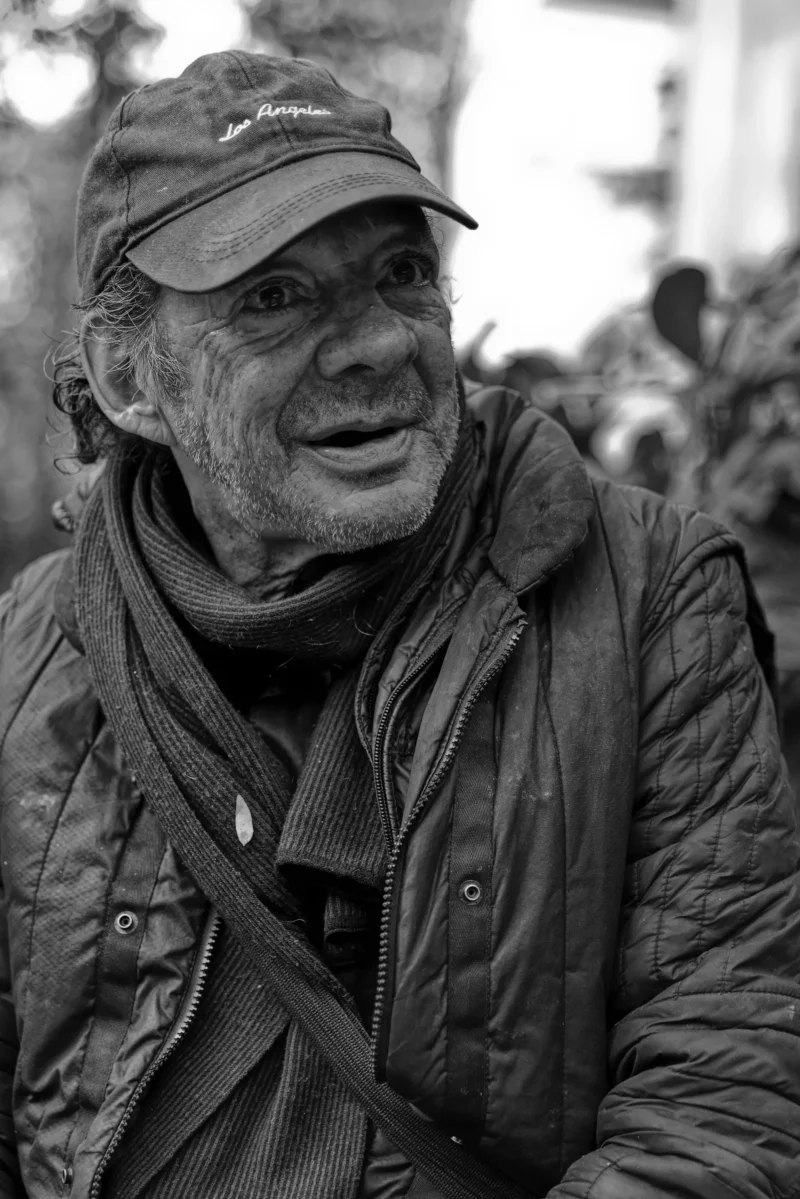 Mickaël · 11/11/2024 · 3519 x 5272 Cette photographie met en scène un homme âgé comme sujet principal, au regard pensif et à l'expression marquée. Le cadrage est serré, centré sur son visage et sa partie supérieure du corps, créant une intimité avec le spectateur.La composition est simple et directe, avec un arrière-plan flou et légèrement déstructuré, mettant l'accent sur le sujet. La lumière est douce et naturelle, sans contraste violent, adoucissant les traits du visage.En noir et blanc, les tons dominants varient du clair au foncé, renforçant les textures et les émotions. Le style est documentaire et réaliste, évoquant une ambiance authentique et mélancolique.