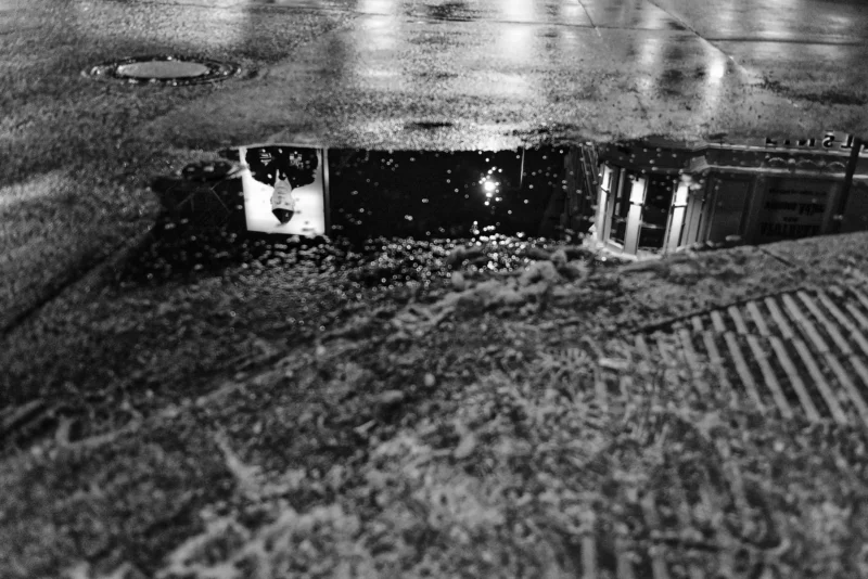 Mickaël · 14/02/2025 · 5096 x 3402 Cette photographie en noir et blanc capture une scène onirique et mystérieuse. Le sujet principal semble être une réflexion dans une flaque d’eau, déformant une structure architecturale, probablement un bâtiment ou un pont.La composition joue sur les symétries et les reflets, créant une ambiance presque surréaliste. Le cadrage est serré, centré sur la flaque et ses reflets. La lumière, tamisée, accentue les contrastes et les textures, renforçant l’aspect dramatique. Les couleurs dominantes sont les nuances de gris, typiques du noir et blanc, ajoutant une touche mélancolique et intemporelle. Le style évoque une esthétique poétique et contemplative.