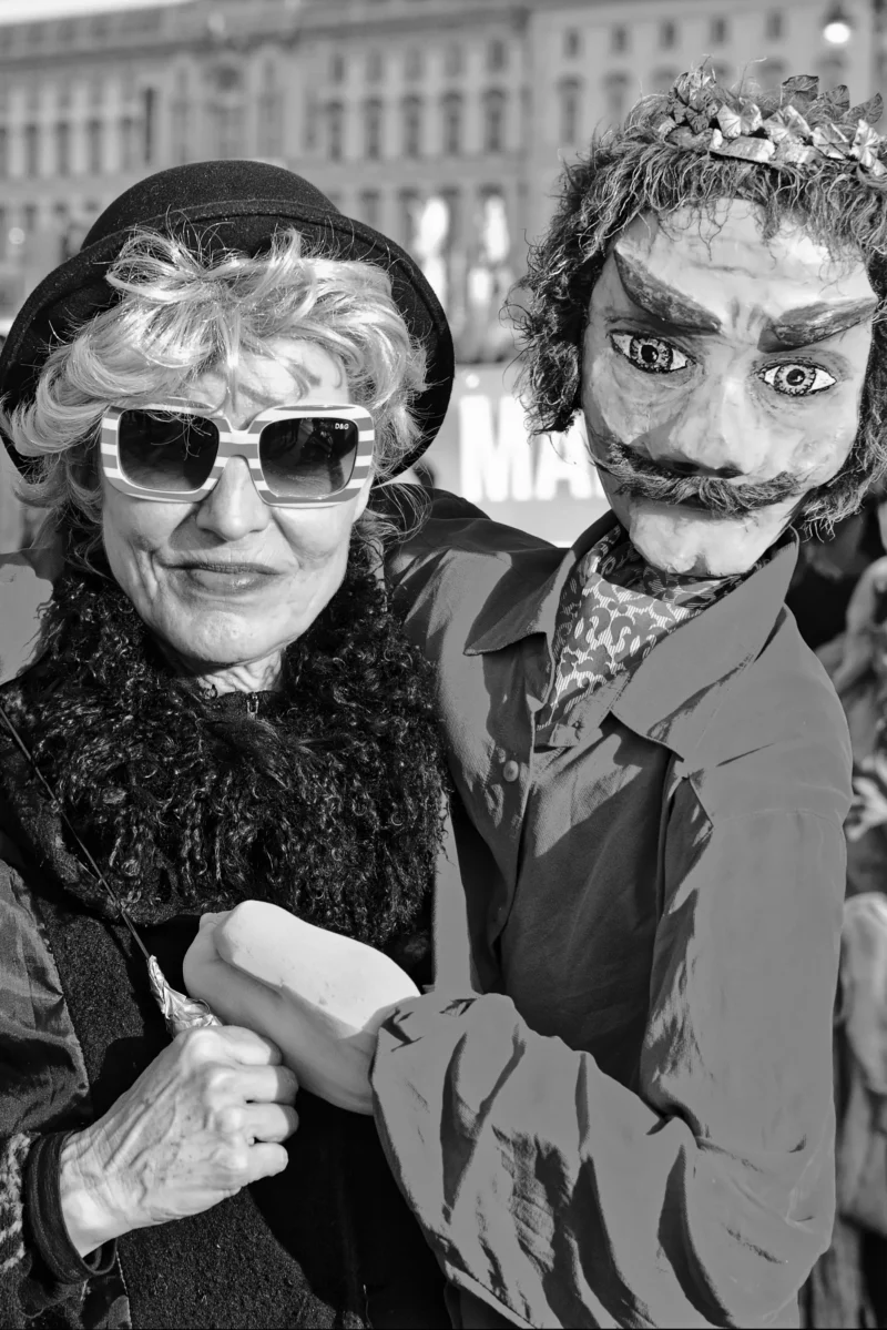 Mickaël · 22/02/2025 · 2678 x 4011 Cette photographie met en scène deux personnes en noir et blanc. Le sujet principal est une femme âgée aux lunettes noires, souriante, accompagnée d’un homme au visage maquillé façon clown ou masque grotesque.La composition est serrée, centrée sur leurs visages et épaules. Le cadrage est rapproché, créant une intimité. La lumière est naturelle, douce, sans contraste marqué. Les couleurs dominantes sont les tons de gris, renforçant l’aspect dramatique et vintage.Le style évoque un mélange de réalisme et de théâtralité, tandis que l’ambiance est à la fois mystérieuse et touchante, presque surréaliste.