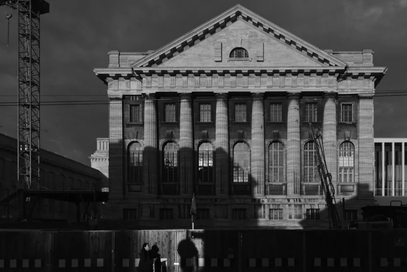 Mickaël · 22/02/2025 · 5477 x 3656 Cette photographie met en scène un bâtiment imposant de style néoclassique, probablement un ancien édifice industriel ou public. Le sujet principal est l’architecture symétrique avec ses colonnes et ses grandes fenêtres.La composition est centrée, avec un cadrage serré qui isole le bâtiment. La lumière, tamisée et contrastée, accentue les textures et les ombres, créant une ambiance dramatique. Les tons dominants sont les gris et noirs, typiques d’un rendu monochrome, renforçant une atmosphère mélancolique et historique. Le style est sobre et classique, évoquant une certaine nostalgie.
