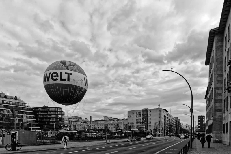 Mickaël · 20/03/2026 · 5550 x 3705 Cette photographie en noir et blanc met en scène un ballon publicitaire géant portant l'inscription "WELT". Le sujet principal est ce ballon suspendu au-dessus d'une rue urbaine.La composition montre une perspective large avec des bâtiments, des tramways et des passants. Le cadrage inclut des éléments architecturaux et des rues animées. La lumière est diffuse, typique d'un jour nuageux, créant des contrastes doux. Les couleurs dominantes, bien que monochromes, suggèrent des tons de gris et noir.Le style est documentaire et urbain, évoquant une ambiance moderne et dynamique dans une ville européenne.