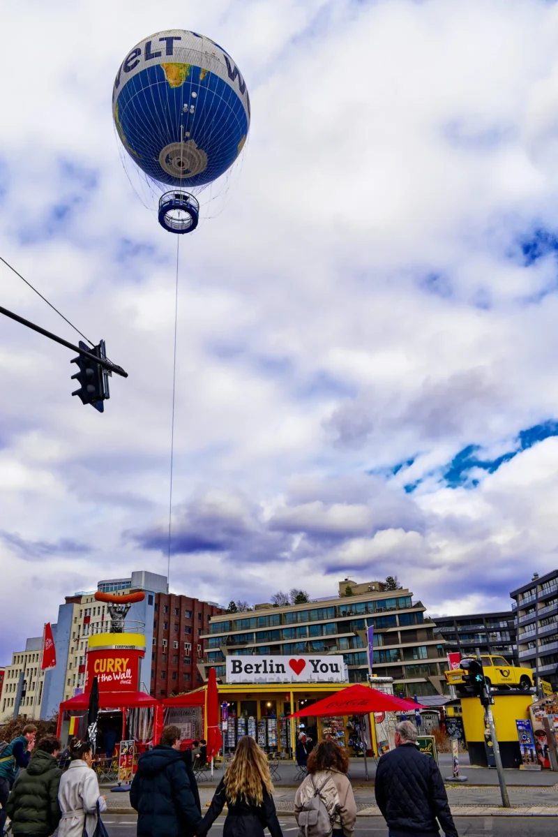 Mickaël · 20/03/2026 · 3814 x 5714 Cette photographie montre une montgolfière en forme de globe comme sujet principal, flottant dans un ciel partiellement nuageux. En bas, une foule se rassemble près d’un stand avec des bannières "Berlin I ♥ Love" et "LT". Le cadrage est dynamique, incluant des éléments urbains et des passants.La lumière est naturelle, avec des contrastes doux. Les couleurs dominantes sont les bleus du ciel, les tons chauds des bâtiments et les touches vives des enseignes rouges et blanches. Le style est documentaire et festif, évoquant une ambiance joyeuse et conviviale, typique d’un événement public en ville.