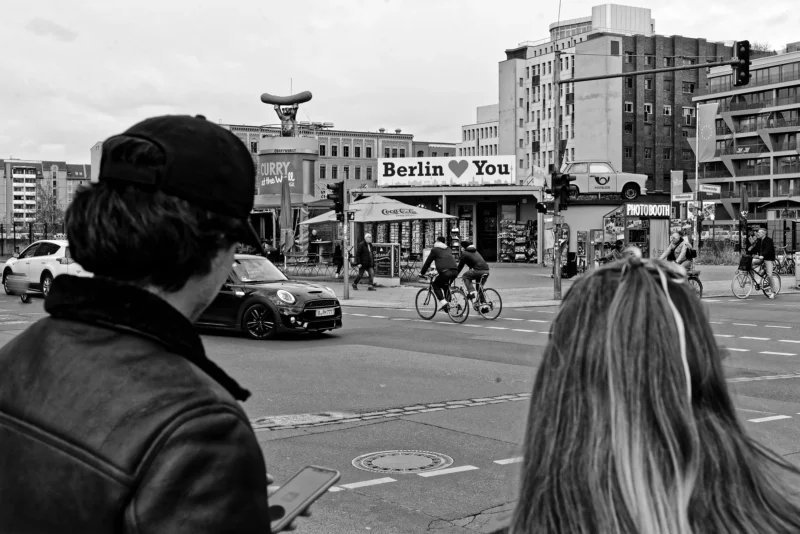 Mickaël · 20/03/2026 · 5503 x 3674 Cette photographie en noir et blanc capture une scène urbaine à Berlin. Le sujet principal est un homme vu de dos, avec une longue chevelure et une veste en cuir. La composition inclut une rue animée avec des voitures, des cyclistes et des bâtiments.Le cadrage est serré sur l’homme, mais inclut aussi l’arrière-plan. La lumière semble naturelle, avec des contrastes marqués. Les couleurs dominantes, bien que monochromes, suggèrent des tons gris et noirs, renforçant l’ambiance urbaine et moderne. Le style est documentaire, capturant un moment authentique. L’ambiance est dynamique et légèrement mélancolique.