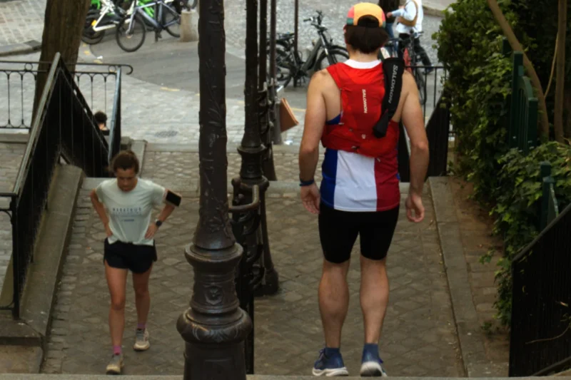 Bao · 1968 x 1312 Cette photographie montre deux personnes en mouvement dans un cadre urbain. Le sujet principal semble être l’homme en premier plan, vêtu d’un tank top rouge et blanc, marchant sur un trottoir en pente.La composition est dynamique, avec un cadrage légèrement bas, mettant en valeur les jambes et le mouvement. La lumière est naturelle, probablement en journée, avec des ombres douces. Les couleurs dominantes sont les tons neutres (gris du trottoir, vert des plantes) et les couleurs vives du vêtement rouge de l’homme.Le style est documentaire et spontané, capturant une scène de vie quotidienne. L’ambiance est active et urbaine.