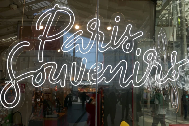 Bao · 23/04/2026 · 5472 x 3648 Cette photographie montre une vitrine avec l'inscription lumineuse "Paris Souvenirs". Le sujet principal est l'écriture néon stylisée, reflétant une ambiance touristique parisienne. La composition met en valeur le texte au premier plan, tandis que le cadrage inclut des éléments flous de l'intérieur, comme des objets et des lumières.La lumière est douce et artificielle, provenant des néons et des éclairages intérieurs. Les couleurs dominantes sont des tons froids (blanc, bleu) avec des touches chaudes reflétées par les lumières internes. Le style est épuré et moderne, évoquant une ambiance à la fois chaleureuse et nostalgique.