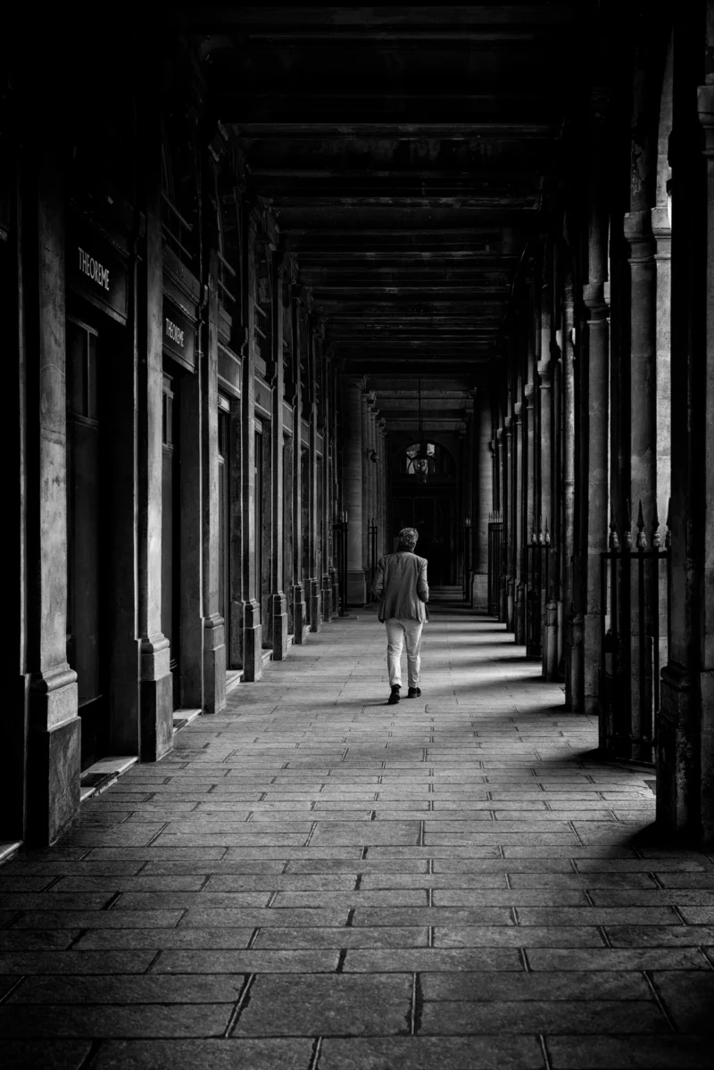 Philippe · 21/02/2026 · 2668 x 4000 Cette photographie en noir et blanc met en scène un homme marchant seul dans une longue allée couverte. Le sujet principal est cet individu, dont la silhouette se détache progressivement dans la profondeur.La composition repose sur une perspective centrale, avec des colonnes et des arches créant un effet de tunnel. Le cadrage serré et la lumière tamisée, provenant probablement d’une source lointaine, accentuent l’atmosphère mystérieuse et contemplative. Les tons dominants, allant du noir au gris clair, renforcent le côté dramatique et introspectif. Le style rappelle le photojournalisme classique ou documentaire, évoquant une ambiance mélancolique et solitaire.