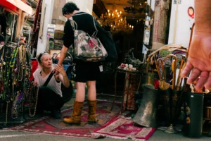 Don · 25/04/2026 · 4672 x 3115 Cette photographie montre une femme accroupie, sujet principal, entourée d’objets artisanaux. Elle porte un sac à dos et des bottes, avec un style décontracté et bohème. La composition est serrée, centrée sur elle et les produits autour (bijoux, tissus, poteries).Le cadrage est dynamique, avec des éléments en profondeur. La lumière est naturelle, légèrement tamisée, créant des ombres douces. Les couleurs dominantes sont chaudes : tons terreux, rouges, bruns et touches de vert. L’ambiance est artisanale, authentique et chaleureuse, évoquant un marché ou une boutique d’artisanat.