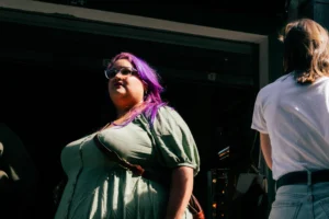 Don · 25/04/2026 · 3906 x 2604 Cette photographie capture une personne au premier plan, vêtue d’une tenue verte et aux cheveux violets, regardant vers la gauche. Le cadrage est serré, mettant en valeur son visage et son expression.L’ambiance semble informelle et artistique, probablement lors d’un événement culturel ou musical. La lumière est naturelle et douce, avec des contrastes marqués créant des ombres nettes. Les couleurs dominantes sont les tons verts, violets et blancs, renforçant une ambiance vibrante et moderne. Le style est spontané et authentique, évoquant une scène intimiste et créative.