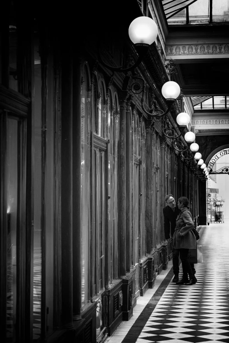 Philippe · 21/02/2026 · 2668 x 4000 Cette photographie en noir et blanc met en scène un homme seul, sujet principal, marchant ou s'arrêtant dans un couloir couvert. La composition est symétrique, avec une perspective guidée par les vitrines alignées et les globes lumineux suspendus. Le cadrage serré et la profondeur de champ accentuent l'isolement du personnage.La lumière douce, tamisée par les lampes, crée une ambiance mélancolique et intemporelle. Les tons gris dominent, renforçant le côté nostalgique et dramatique. Le style rappelle le photojournalisme classique ou documentaire, évoquant une atmosphère à la fois urbaine et contemplative.