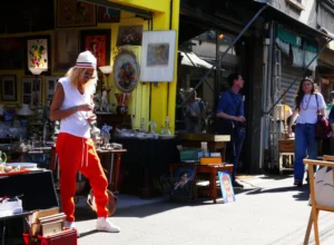 Bao · 4302 x 3152 Cette photographie capture une scène de rue animée. Le sujet principal est un artiste de rue, vêtu d’un costume blanc et d’un pantalon rouge, en train de jouer de la guitare. La composition montre un marché artisanal en arrière-plan avec des étals colorés et des objets variés. Le cadrage est large, incluant plusieurs personnes et éléments urbains.La lumière est naturelle et éclatante, suggérant une journée ensoleillée. Les couleurs dominantes sont vives : jaune, rouge, blanc et des teintes chaudes. Le style est documentaire et spontané, évoquant une ambiance festive et artistique dans un cadre urbain.