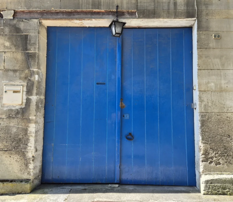 Bao · 3446 x 3000 Cette photographie montre une porte industrielle bleue en métal, sujet principal. La composition est simple et centrée, avec un cadrage serré sur la porte, encadrée par un mur en béton gris.La lumière semble naturelle, mais diffuse, sans ombres marquées. Les couleurs dominantes sont le bleu intense de la porte et le gris du béton. Le style est réaliste et documentaire, sans retouches apparentes. L’ambiance est sobre et industrielle, évoquant un lieu utilitaire et peu fréquenté.