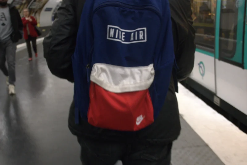 Bao · 5488 x 3664 Cette photographie montre une personne vue de dos, portant un sac à dos bleu avec une bande blanche et rouge. Le sujet principal est donc le sac, marqué du mot "LIEUX".La composition est simple, centrée sur le sac. Le cadrage est serré, mettant en valeur le détail du sac et une partie de l’environnement urbain (sol et murs). La lumière semble naturelle et diffuse, typique d’un intérieur éclairé artificiellement.Les couleurs dominantes sont le bleu, le blanc et le rouge du sac, contrastant avec les tons neutres du fond. L’ambiance est sobre et quotidienne, évoquant un lieu public comme une gare ou un centre commercial.