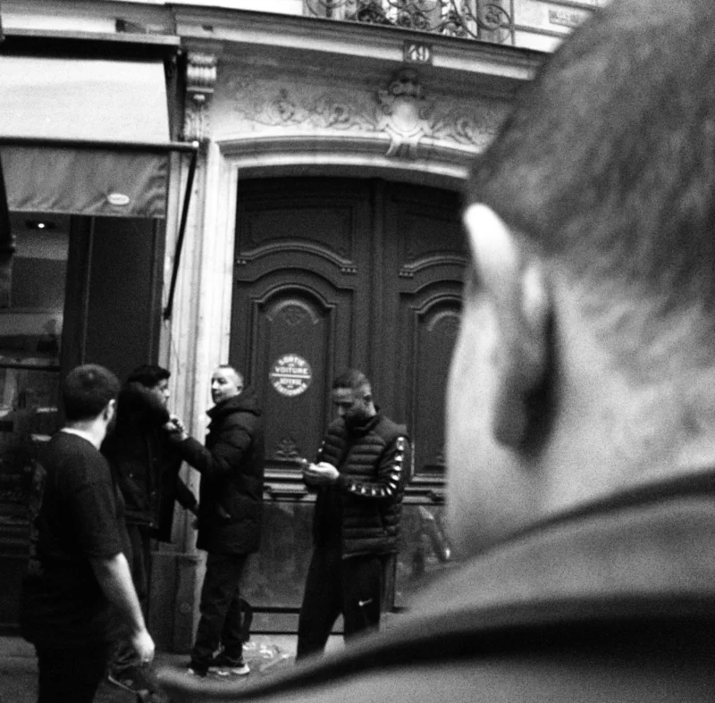 Bao · 4080 x 4016 Cette photographie en noir et blanc capture une scène urbaine animée. Le sujet principal semble être un groupe de personnes devant une porte décorée, dont une figure centrale, probablement un artiste de rue, semble interagir avec le public. La composition est serrée, avec un cadrage qui met l’accent sur les visages et les interactions. La lumière est naturelle, légèrement contrastée, créant des ombres marquées. L’ambiance est dynamique et spontanée, évoquant une atmosphère de rue vivante et artistique, typique d’un spectacle improvisé. Le style est documentaire, authentique et immersif.
