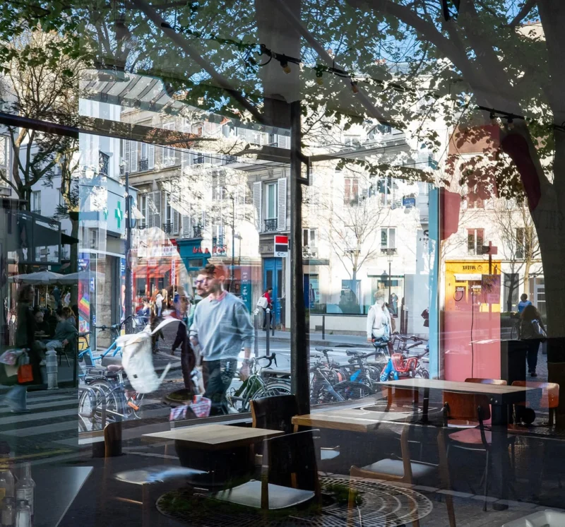 Nathalie C H · 04/04/2026 · 1333 x 1243 Cette photographie montre une vue intérieure depuis un café ou un restaurant. Le sujet principal est la rue animée visible à travers les grandes vitres.La composition joue sur la transparence entre intérieur et extérieur, avec des tables vides en premier plan. Le cadrage est large, capturant une partie de la rue et des bâtiments colorés. La lumière est naturelle, éclatante, créant des contrastes et des ombres douces. Les couleurs dominantes sont vives : tons pastel, bleus, roses et verts.Le style est urbain et contemporain, évoquant une ambiance dynamique et chaleureuse, typique d’une ville européenne.