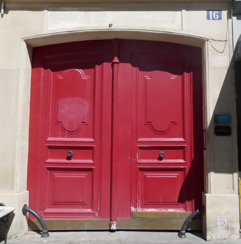 Bao · 3596 x 3648 Cette photographie montre une **porte d'entrée rouge** comme sujet principal. La composition est centrée sur cette porte en bois, aux panneaux décoratifs et poignées noires. Le cadrage est serré, mettant en valeur les détails architecturaux.La lumière semble naturelle, éclairant uniformément la porte. Les **couleurs dominantes** sont le rouge vif de la porte et le beige clair du mur. Le style évoque un charme classique et traditionnel, typique des anciennes maisons européennes. L’ambiance est à la fois chaleureuse et élégante, renforcée par les contrastes entre les matériaux et les couleurs.