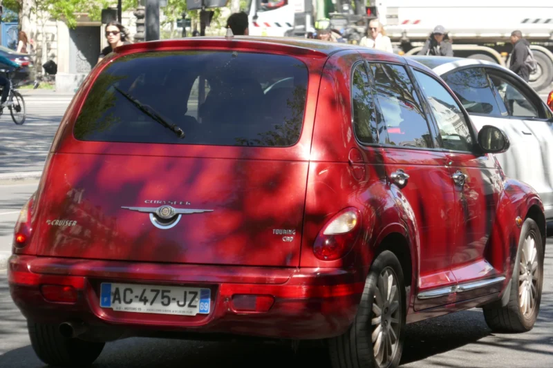 Bao · 23/04/2026 · 5472 x 3648 Cette photographie met en scène une **Peugeot 206** rouge, sujet principal, dont la carrosserie est partiellement endommagée. Le **cadrage** est serré sur l’arrière du véhicule, en position statique dans un environnement urbain.La **composition** inclut des éléments flous en arrière-plan, comme des passants et des arbres. La **lumière** semble naturelle, avec des ombres douces, suggérant un jour ensoleillé. Les **couleurs dominantes** sont le rouge vif de la voiture et les tons neutres de l’asphalte et des vêtements des passants.L’ambiance est **urbaine et réaliste**, avec une touche de **décadence** due aux dommages visibles sur la voiture.