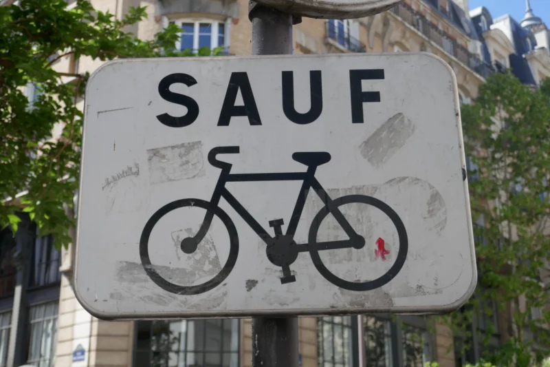 Bao · 23/04/2026 · 5472 x 3648 Cette photographie montre un panneau de signalisation urbaine avec le mot **« SAUF »** en haut et un dessin de vélo barré en dessous. Le sujet principal est ce panneau, légèrement abîmé et graffité.La composition est simple, centrée sur le panneau, avec un cadrage serré. L’arrière-plan montre un bâtiment et des arbres, suggérant une rue en ville. La lumière est naturelle, probablement en journée, avec des ombres douces. Les couleurs dominantes sont les tons gris (panneau), vert (végétation) et beige (bâtiment).Le style est réaliste et documentaire, évoquant une ambiance urbaine et quotidienne.