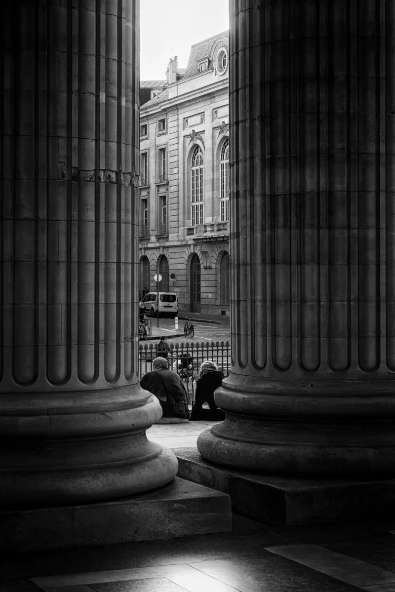 Philippe · 08/11/2025 · 2668 x 4000 Cette photographie en noir et blanc capture un espace architectural imposant. Le sujet principal est l'alignement des colonnes massives encadrant la vue. La composition joue sur la perspective avec un cadrage serré entre les piliers, créant une profondeur vers un bâtiment classique en arrière-plan.La lumière, tamisée et contrastée, accentue les ombres et les textures. Les couleurs dominantes, bien que monochromes, exploitent les nuances de gris pour renforcer le dramatique. Le style est classique et intemporel, évoquant une ambiance solennelle et mystérieuse, presque théâtrale.