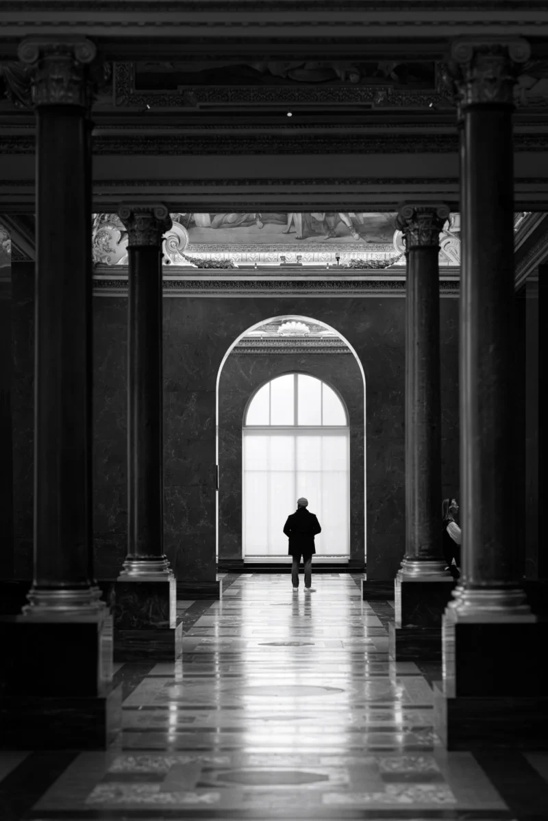 Philippe · 19/11/2025 · 2001 x 3000 Cette photographie met en scène un **sujet principal** : une silhouette humaine en mouvement dans un corridor. La **composition** repose sur une perspective centrale, avec des colonnes et des arches symétriques encadrant le passage. Le **cadrage** est serré, guidant le regard vers le fond.La **lumière**, naturelle et tamisée, crée des contrastes forts, accentuant les ombres et les volumes. Les **couleurs dominantes** sont en noir et blanc, renforçant l'atmosphère dramatique.Le **style** est épuré et classique, évoquant une ambiance **solennelle et mystérieuse**, presque cinématographique.