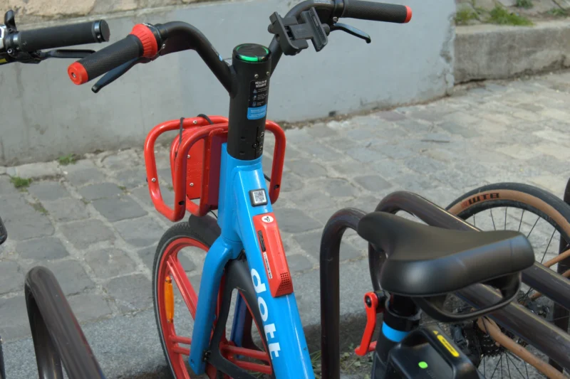 Bao · 6024 x 4016 Cette photographie montre principalement un vélo électrique bleu et rouge garé près d’un autre vélo classique noir. La composition met en avant les détails techniques et colorés du vélo électrique, avec un cadrage serré sur les poignées et le guidon.La lumière semble naturelle, douce et légèrement tamisée, créant des ombres douces. Les couleurs dominantes sont le bleu vif, le rouge et le noir, apportant une touche dynamique et moderne. Le style est simple et réaliste, reflétant un cadre urbain. L’ambiance est à la fois pratique et esthétique, évoquant un environnement urbain contemporain.