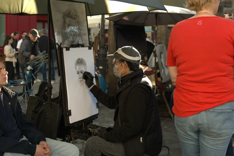 Bao · 6024 x 4016 Cette photographie montre un artiste en train de peindre un portrait en public. Le sujet principal est le peintre, assis et concentré sur son œuvre. La composition est dynamique, avec plusieurs personnes autour, dont certaines observent.Le cadrage est serré sur l’artiste et son chevalet, intégrant partiellement les spectateurs. La lumière semble naturelle, bien que légèrement tamisée, créant des contrastes doux. Les couleurs dominantes sont neutres (noirs, gris, blancs) avec des touches de rouge et de bleu dans les vêtements des spectateurs.L’ambiance est vivante et artistique, typique d’un événement public ou d’un marché.