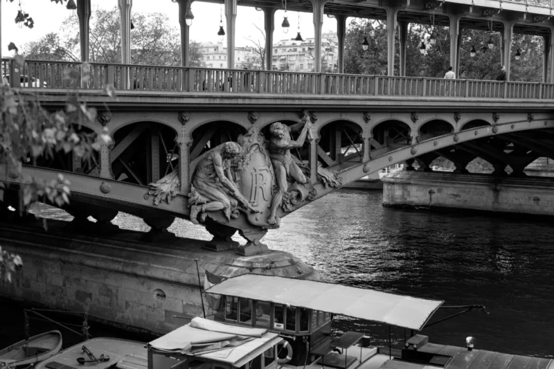 Bao · 2800 x 1867 Cette photographie en noir et blanc montre un **pont métallique** enjambant un canal, avec des **piliers architecturaux** ornés. Le **sujet principal** est le pont et les bateaux amarrés en contrebas.La **composition** est symétrique, avec une perspective centrée. Le **cadrage** est serré, mettant en valeur les détails des structures. La **lumière**, douce et diffuse, crée des contrastes modérés. Les **couleurs dominantes** sont les nuances de gris, renforçant l’aspect vintage.Le **style** est documentaire, évoquant une ambiance **nostalgique et industrielle**, typique des scènes urbaines anciennes.