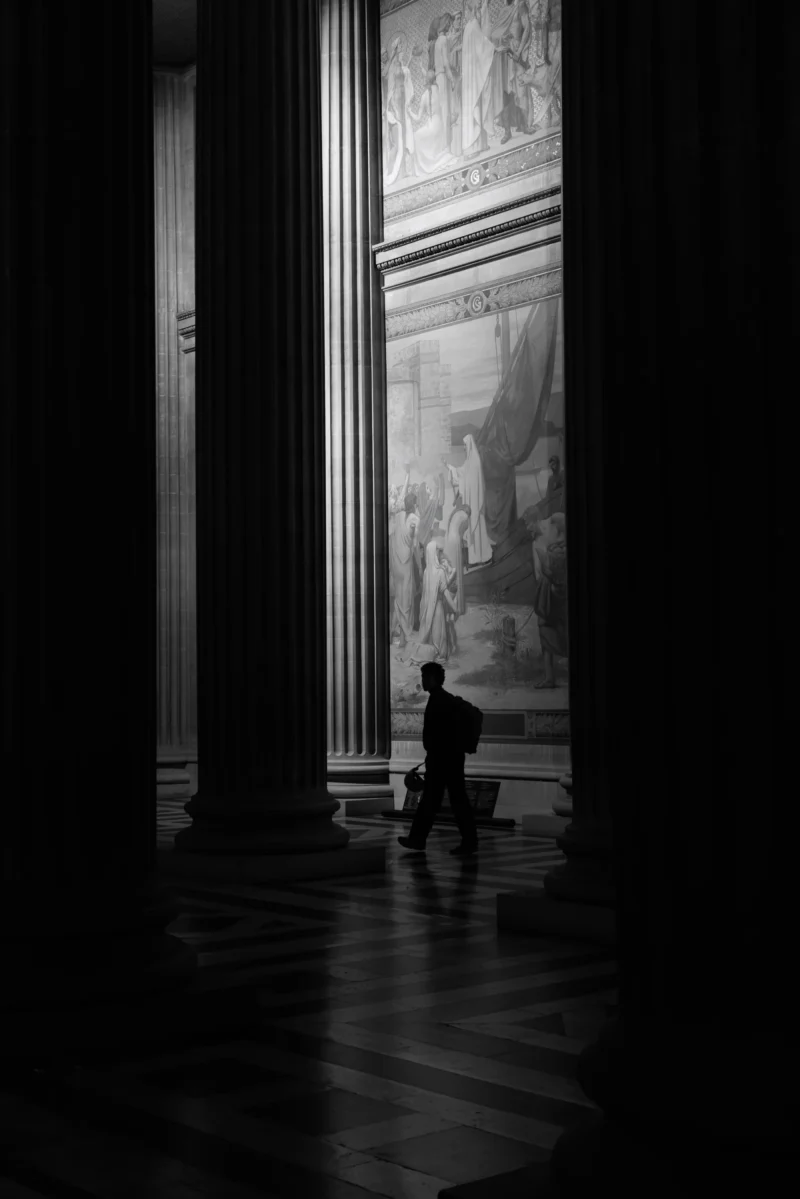 Philippe · 2001 x 3000 Cette photographie en noir et blanc met en scène un homme assis seul au sol, dans une vaste pièce architecturale imposante. Le sujet principal est isolé, créant une atmosphère contemplative. La composition joue sur les lignes verticales des colonnes et l’ouverture lumineuse en arrière-plan, renforçant la profondeur et la symétrie. Le cadrage serré et la lumière naturelle filtrée accentuent les contrastes, soulignant les ombres et les volumes. Les tons dominants, du blanc au noir, renforcent le côté dramatique et intemporel. Le style évoque une ambiance mélancolique et solennelle, typique des espaces historiques ou sacrés.