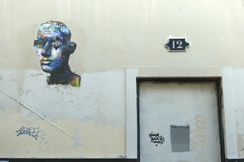 Bao · 6024 x 4016 Cette photographie montre un mural représentant un buste stylisé avec des traits graphiques et des couleurs vives. Le sujet principal est ce portrait, situé à gauche de la porte numérotée 12.La composition est simple, avec un cadrage serré sur le mural et la porte. La lumière naturelle éclaire uniformément la scène, sans ombres marquées. Les couleurs dominantes sont des tons froids (beige de la façade) contrastant avec les touches chaudes et colorées du graffiti (bleu, vert, rouge).Le style est urbain et artistique, évoquant une ambiance street art, dynamique et créative.