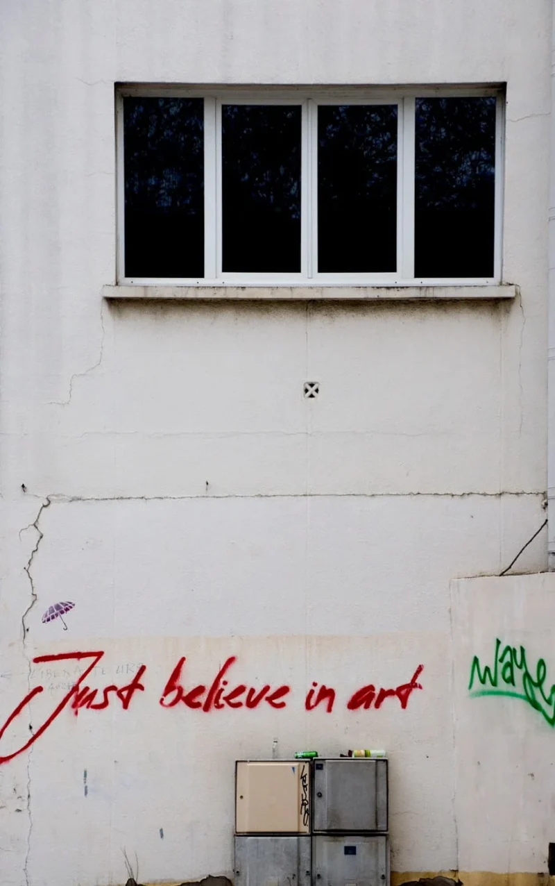 Franck T. · 16/01/2026 · 878 x 1400 Cette photographie met en scène un mur blanc dégradé avec une inscription en rouge : *« Just believe in art »*. Le sujet principal est cette phrase graffitée, soulignée par un trait de couleur vive.La composition est épurée, centrée sur le texte et les trois fenêtres grillagées au-dessus. Le cadrage est serré, mettant en valeur les détails du mur et de l’écriture. La lumière naturelle éclaire uniformément la scène, créant des contrastes doux.Les couleurs dominantes sont le blanc du mur, le rouge vif du graffiti et les teintes grises des fenêtres. Le style est urbain et brut, évoquant une ambiance à la fois minimaliste et inspirante, mêlant simplicité et message artistique.