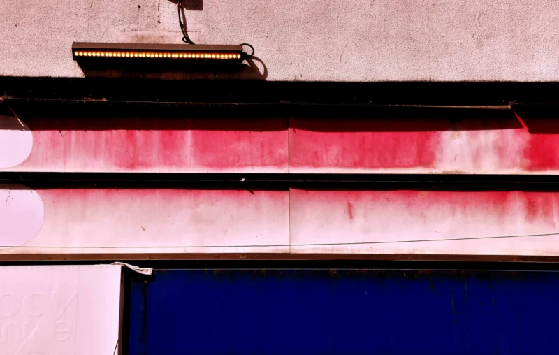 Franck T. · 20/04/2026 · 5490 x 3485 Cette photographie met en scène un détail architectural minimaliste. Le sujet principal est une enseigne métallique usée, légèrement rouillée, fixée à une façade de bâtiment.La composition repose sur des lignes horizontales et verticales, avec un cadrage serré qui isole ces éléments. La lumière, douce et naturelle, semble provenir d’un côté, créant des contrastes subtils.Les couleurs dominantes sont le rouge (teinte de la façade), le bleu profond (porte ou fenêtre) et les tons métalliques (enseigne). Le style est épuré et documentaire, évoquant une ambiance urbaine et légèrement mélancolique, avec une touche d’industrialisation.