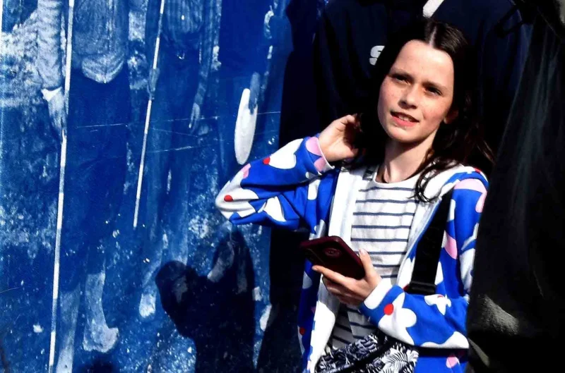 Franck T. · 15/04/2026 · 1400 x 926 Cette photographie met en scène une jeune femme comme sujet principal, posant contre un mur aux carreaux bleus. Elle porte un pull à rayures horizontales et une veste bleue à motifs. Le cadrage est serré, centré sur elle, avec une légère inclinaison.La lumière semble naturelle et douce, créant des ombres douces. Les couleurs dominantes sont les bleus froids du mur et les tons neutres de ses vêtements. Le style est simple et spontané, évoquant une ambiance intime et légèrement mélancolique.