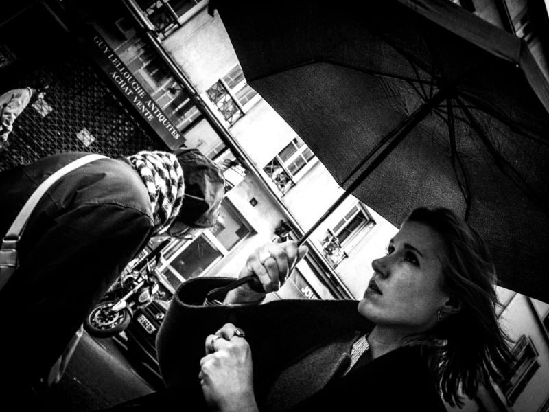 Jean-Philippe · 11/04/2026 · 7304 x 4869 Cette photographie en noir et blanc met en scène une jeune femme comme sujet principal, assise sous un parapluie, semblant absorbée par ses pensées. Le cadrage est serré sur son visage, avec une légère inclinaison vers le haut, créant une impression d’intimité.La composition joue sur les contrastes entre l’ombre et la lumière, accentués par le parapluie ouvert. Les tons dominants sont les gris et noirs, renforçant une ambiance mélancolique et contemplative. Le style est documentaire et poétique, évoquant une atmosphère introspective et urbaine.
