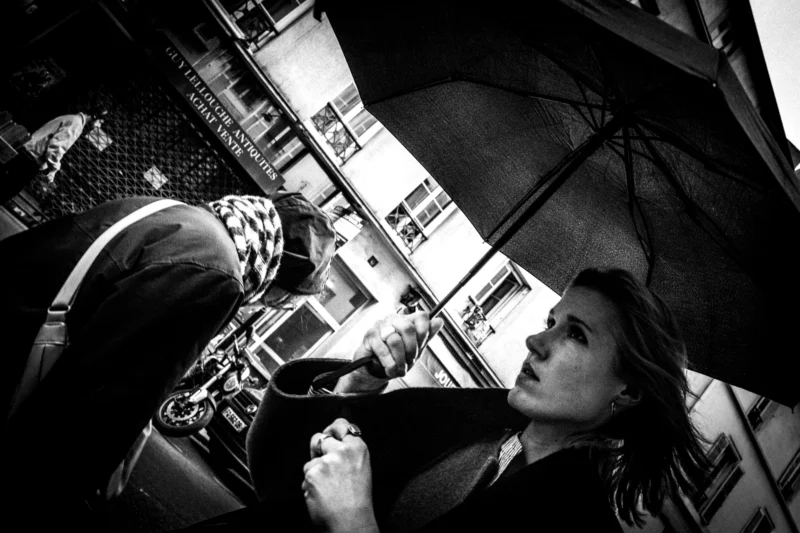 Jean-Philippe · 11/04/2026 · 7304 x 4869 Cette photographie en noir et blanc met en scène une jeune femme comme sujet principal, assise sous un parapluie, semblant absorbée par ses pensées. Le cadrage est serré sur son visage, avec une légère inclinaison vers le haut, créant une impression d’intimité.La composition joue sur les contrastes entre l’ombre et la lumière, accentués par le parapluie ouvert. Les tons dominants sont les gris et noirs, renforçant une ambiance mélancolique et contemplative. Le style est documentaire et poétique, évoquant une atmosphère introspective et urbaine.