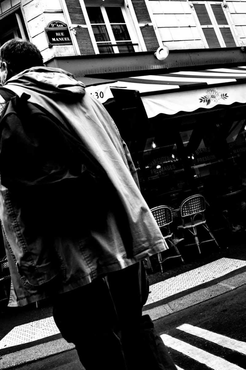 Jean-Philippe · 11/04/2026 · 5424 x 8136 Cette photographie en noir et blanc met en scène un homme de dos, vêtu d’un manteau et d’un sac à dos, marchant dans une rue. Le sujet principal est flou, attirant l’attention sur son mouvement et son attitude.La composition est simple et épurée, avec un cadrage serré sur le personnage. L’arrière-plan montre des tables et des numéros de rue, suggérant un cadre urbain. La lumière est douce, créant des contrastes subtils. Le style est documentaire et introspectif, renforcé par le monochrome.L’ambiance est calme et mélancolique, évoquant une solitude urbaine.
