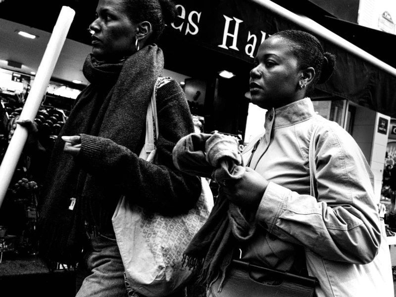 Jean-Philippe · 11/04/2026 · 7141 x 5356 Cette photographie en noir et blanc met en scène deux personnes comme sujets principaux, dont l’une semble ajuster un accessoire de l’autre. La composition est serrée, centrée sur leurs interactions. Le cadrage est rapproché, capturant des détails des vêtements et des expressions.La lumière est contrastée, avec des ombres marquées, typique d’un éclairage urbain. Les couleurs dominantes, bien que monochromes, jouent sur les nuances de gris.Le style est documentaire et spontané, évoquant une ambiance authentique et intime, presque théâtrale. L’image dégage une touche de mélancolie et de modernité.