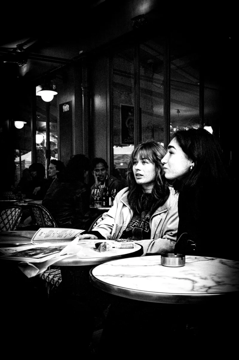 Jean-Philippe · 11/04/2026 · 3816 x 5750 Cette photographie en noir et blanc met en scène deux jeunes femmes assises dans un café. Le sujet principal est leur échange intimiste, capturé de profil.La composition est simple et centrée, avec un cadrage serré sur les deux personnages. L'arrière-plan flou suggère un intérieur chaleureux, probablement un restaurant ou un café.La lumière douce et tamisée crée une ambiance intimiste et nostalgique. Les contrastes accentuent les détails des visages et des objets sur la table.Le style rappelle le photojournalisme classique, évoquant une scène de vie quotidienne. L'absence de couleurs renforce l'aspect intemporel et émotionnel de l'image.