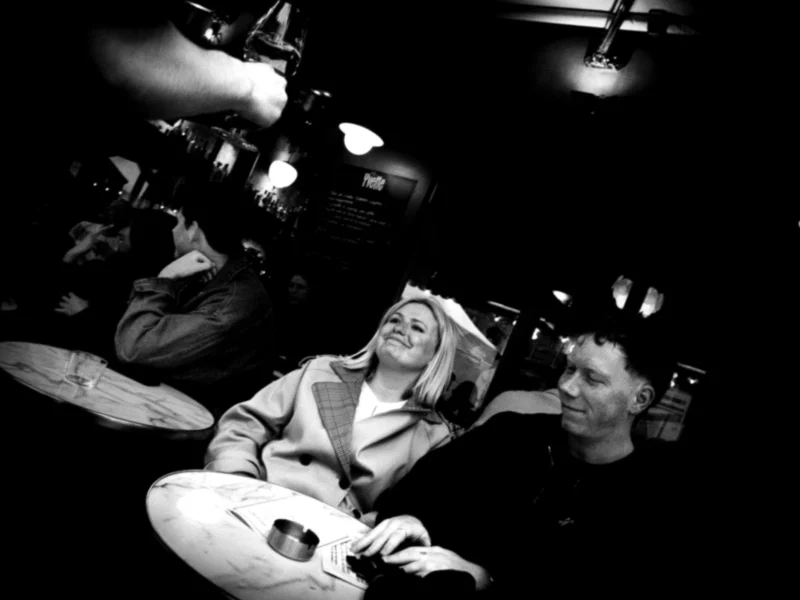 Jean-Philippe · 11/04/2026 · 8091 x 6068 Cette photographie en noir et blanc met en scène deux personnes assises dans un espace confiné, probablement un véhicule. Le sujet principal semble être la femme allongée, vêtue d’un manteau clair, avec un homme à ses côtés.La composition est serrée, avec un cadrage rapproché qui crée une intimité. Les lumières sont tamisées, avec des éclairages contrastés, donnant une ambiance sombre et mystérieuse. Les couleurs dominantes, bien que monochromes, jouent sur les nuances de gris et de noir, renforçant l’atmosphère dramatique et mélancolique. Le style rappelle un documentaire ou une scène cinématographique réaliste.