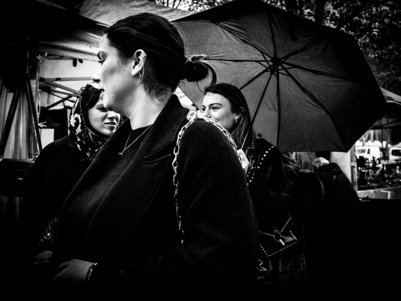 Jean-Philippe · 11/04/2026 · 6784 x 5088 Cette photographie en noir et blanc met en scène deux femmes comme sujets principaux. La composition montre une femme de profil, vêtue d’un manteau sombre, et une autre partiellement visible sous un parapluie.Le cadrage est serré, centré sur elles, avec un arrière-plan flou. La lumière est douce, créant des contrastes subtils et des ombres marquées. Les couleurs dominantes sont les nuances de gris, renforçant l’aspect dramatique et intemporel.Le style évoque une ambiance rétro, presque cinématographique, avec une touche mélancolique et contemplative. L’atmosphère est calme et introspective.