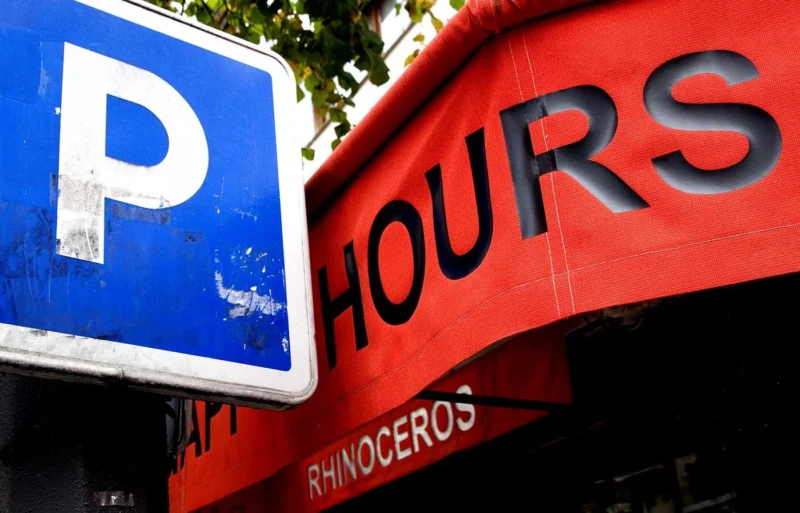 Franck T. · 22/10/2025 · 1400 x 897 Cette photographie met en avant deux panneaux superposés. Le sujet principal est un panneau rouge indiquant "HOURS" (horaires) partiellement caché par un panneau bleu de stationnement "P".La composition est serrée, avec un cadrage rapproché. La lumière semble naturelle, bien que légèrement contrastée. Les couleurs dominantes sont le bleu vif du panneau de stationnement et le rouge du panneau d'horaires, avec des touches de vert en arrière-plan.Le style est simple et direct, évoquant une ambiance urbaine et un peu désordonnée. L'image suggère un lieu commercial ou un parking.