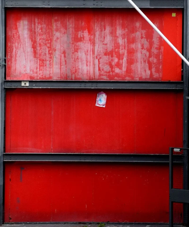 Franck T. · 22/09/2025 · 836 x 1000 Cette photographie montre un conteneur ou une porte métallique rouge écaillée et abîmée. Le sujet principal est la surface rouillée et dégradée, avec des traces de corrosion et de chocs.La composition est simple et centrée, avec un cadrage serré qui isole presque entièrement le panneau rouge. Les couleurs dominantes sont le rouge vif et le noir des bordures métalliques. La lumière semble naturelle mais diffuse, mettant en valeur les imperfections.Le style est réaliste et brut, évoquant une ambiance industrielle et usée, presque mélancolique.