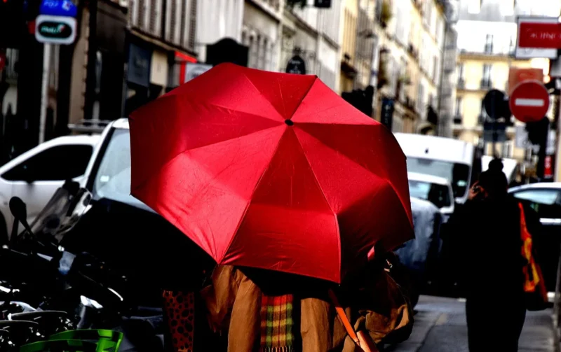 Franck T. · 12/02/2026 · 1400 x 880 Cette photographie met en scène un **parapluie rouge** comme sujet principal, ouvert et dominant l'image. La composition montre une rue urbaine avec des éléments flous en arrière-plan, comme des voitures et des passants.Le **cadrage** est serré sur le parapluie, isolant partiellement le sujet. La **lumière** semble naturelle mais tamisée, créant des contrastes doux. Les **couleurs dominantes** sont le rouge vif du parapluie et des tons neutres (gris, beige) pour le reste.Le style est **urbain et réaliste**, avec une ambiance légèrement floue et dynamique, évoquant une atmosphère **vivante et mouvementée**.