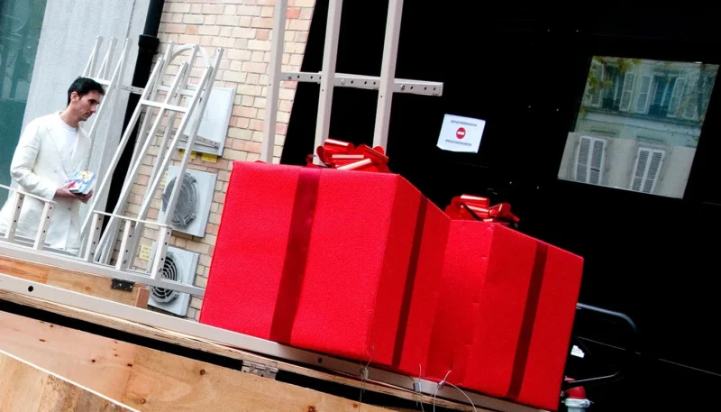 Franck T. · 01/12/2025 · 1400 x 800 Cette photographie montre comme sujet principal trois grandes valises rouges posées sur une structure en bois, évoquant un mobilier ou une installation artistique. En arrière-plan, un homme est partiellement visible, ainsi qu’un cadre architectural avec des escaliers et des murs en brique.La composition est symétrique et géométrique, avec un cadrage serré sur les valises. La lumière semble naturelle, mais contrastée, créant des ombres marquées. Les couleurs dominantes sont le rouge vif des valises et les tons neutres des matériaux (bois, brique). Le style semble contemporain, avec une ambiance à la fois industrielle et artistique, évoquant une scène de stockage ou une œuvre conceptuelle.