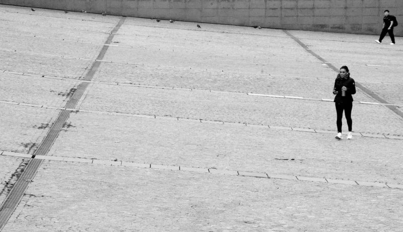 Franck T. · 08/12/2025 · 1400 x 809 Cette photographie en noir et blanc met en scène une personne comme sujet principal, debout au centre d’une vaste place vide et carrelée. La composition est épurée, avec une perspective symétrique et des lignes droites convergeant vers l’horizon.Le cadrage est large, capturant l’immensité de l’espace. La lumière, naturelle et douce, crée des contrastes subtils entre ombres et clartés. Les couleurs dominantes, bien que monochromes, jouent sur les nuances de gris.Le style est sobre et minimaliste, évoquant une ambiance calme et presque mélancolique, renforcée par l’isolement du personnage dans ce cadre architectural.