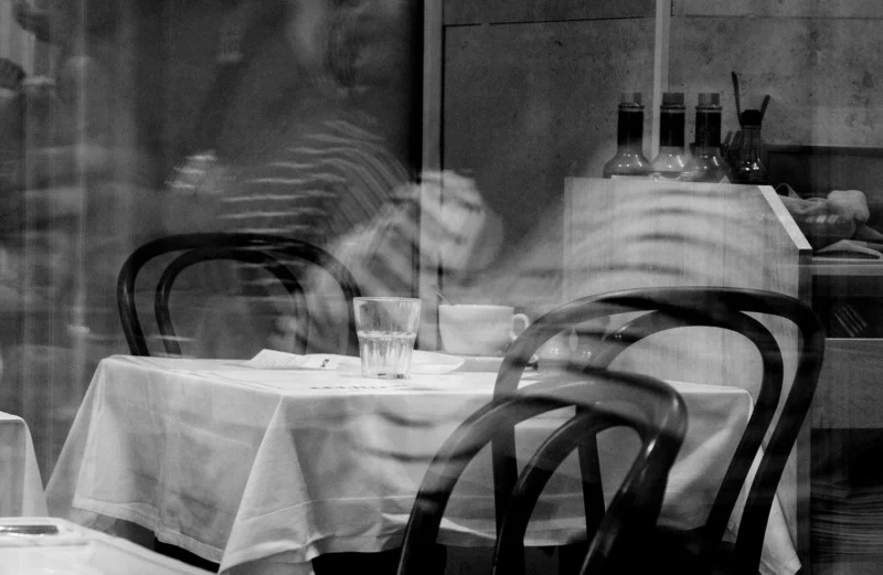 Pascale · 01/05/2026 · 1500 x 977 Cette photographie en noir et blanc montre un **restaurant peu fréquenté**. Le **sujet principal** est une table dressée avec des couverts et une nappe, partiellement floue, évoquant une atmosphère déserte.La **composition** met en valeur un espace vide, avec des chaises et une table bien arrangée. Le **cadrage** est serré, centré sur la table et légèrement incliné. La **lumière** est douce et tamisée, créant des contrastes subtils.Les **couleurs dominantes** sont les tons de gris, renforçant une ambiance **nostalgique et mélancolique**. Le style est **minimaliste et réaliste**, évoquant une scène calme et presque abandonnée.