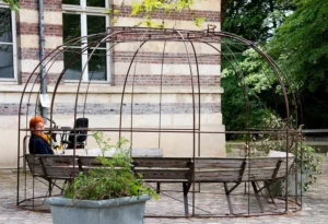 Pascale · 03/05/2026 · 1500 x 1025 Cette photographie met en scène un **sujet principal** : un homme assis sur un banc sous une structure métallique en forme d'arche, entourée de plantes.La **composition** est équilibrée, avec un cadrage serré sur le banc et ses occupants, encadré par les arches métalliques. La **lumière** est naturelle, douce et légèrement tamisée, créant des ombres douces.Les **couleurs dominantes** sont les tons verts des plantes, les gris métalliques des arches, et les teintes neutres du banc et des murs. Le **style** est réaliste et documentaire.L’**ambiance** est calme et introspective, évoquant la sérénité d’un espace urbain végétalisé.
