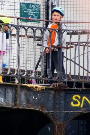 Pascale · 03/05/2026 · 1005 x 1500 Cette photographie montre un enfant en tenue de travail (gilet orange, casquette) debout sur une structure métallique rouillée, probablement un véhicule ou un engin de chantier. Le sujet principal est l’enfant, cadré en plan serré, avec un arrière-plan flou de grillage et de structures industrielles.La lumière semble naturelle, bien que contrastée, mettant en valeur les détails de la tenue et de la structure. Les couleurs dominantes sont les tons gris métalliques, l’orange vif du gilet et les touches de vert et bleu en arrière-plan. Le style est documentaire, capturant une scène réaliste et spontanée, évoquant une ambiance à la fois industrielle et touchante.