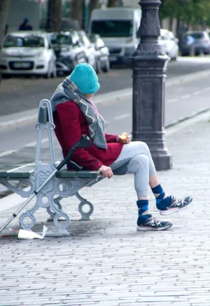 Pascale · 03/05/2026 · 1033 x 1500 Cette photographie montre une personne assise sur un banc en forme de vélo, sujet principal. Elle porte un casque bleu, un manteau rouge et des bottes, tenant un objet à la main.La composition est simple, avec un cadrage serré sur le sujet, mettant en valeur son activité insolite. L’arrière-plan montre une rue urbaine avec des voitures et des arbres.La lumière est naturelle, probablement en journée, avec des ombres douces. Les couleurs dominantes sont les tons rouges, bleus et gris. Le style est documentaire et réaliste, créant une ambiance à la fois urbaine et décalée, mêlant quotidien et originalité.