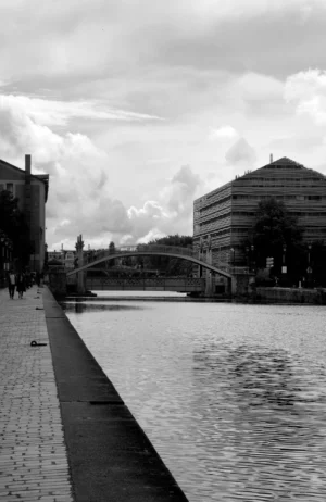 Pascale · 03/05/2026 · 974 x 1500 Cette photographie en noir et blanc met en scène un canal urbain comme sujet principal. La composition montre un pont enjambant l’eau, avec des bâtiments modernes et historiques en arrière-plan. Le cadrage est serré sur le bord du canal, guidant le regard vers le pont et les constructions.La lumière est douce, avec des contrastes modérés, créant une ambiance calme et mélancolique. Les tons dominants sont les gris et noirs, accentués par les reflets sur l’eau. Le style est documentaire et sobre, évoquant une atmosphère paisible et introspective.